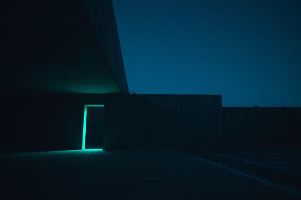 A dark concrete building at night under a deep blue sky, one doorway glowing vivid teal, its light pooling on the ground.